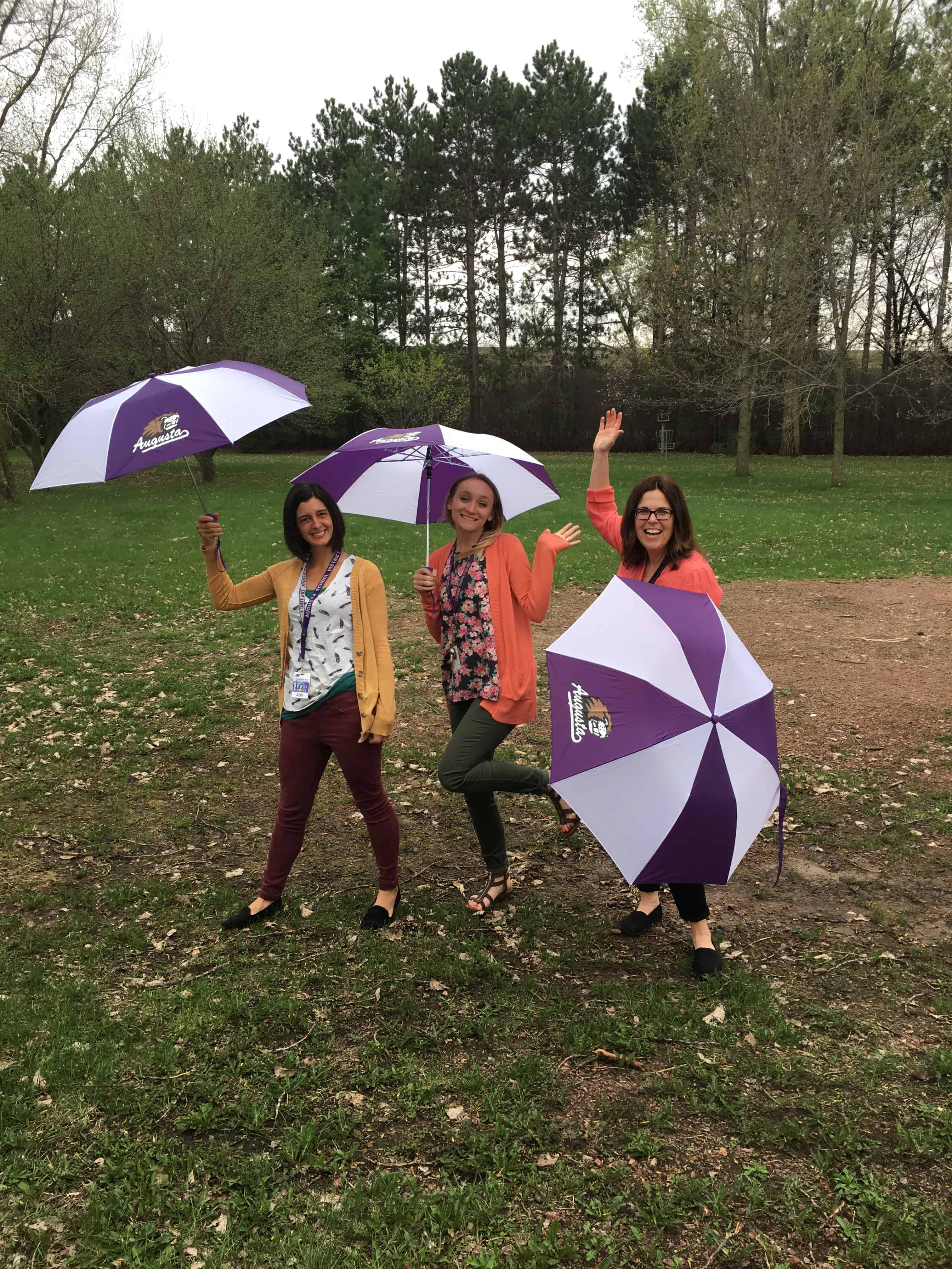 Three staff members laughing and posing outside with their white and purple umbrellas