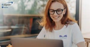 Red haired woman looking at laptop computer. She is wearing glasses and T-shirt with a logo.