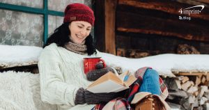 Person bundled up in hat and blanket reading a book outdoors in a winter cabin setting.