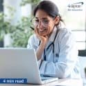 Smiling doctor with white coat and stethoscope looking into laptop.