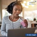 A young woman on her laptop and wearing headphones smiles as she sips a berry smoothie.