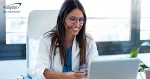 Woman wearing stethoscope and white coat looking at computer monitor and smiling.