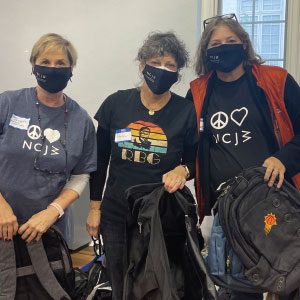 Three women wearing face masks with a logo and holding branded backpacks.