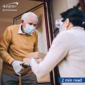 volunteer in protective mask and gloves handing package to elderly man wearing a mask at his front door