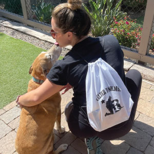 Woman kneeling with her arm around a large dog. She is wearing a drawstring bag imprinted with a soldier and a dog.