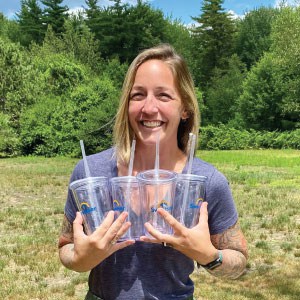 Smiling woman outside holding four clear branded tumblers with straws.
