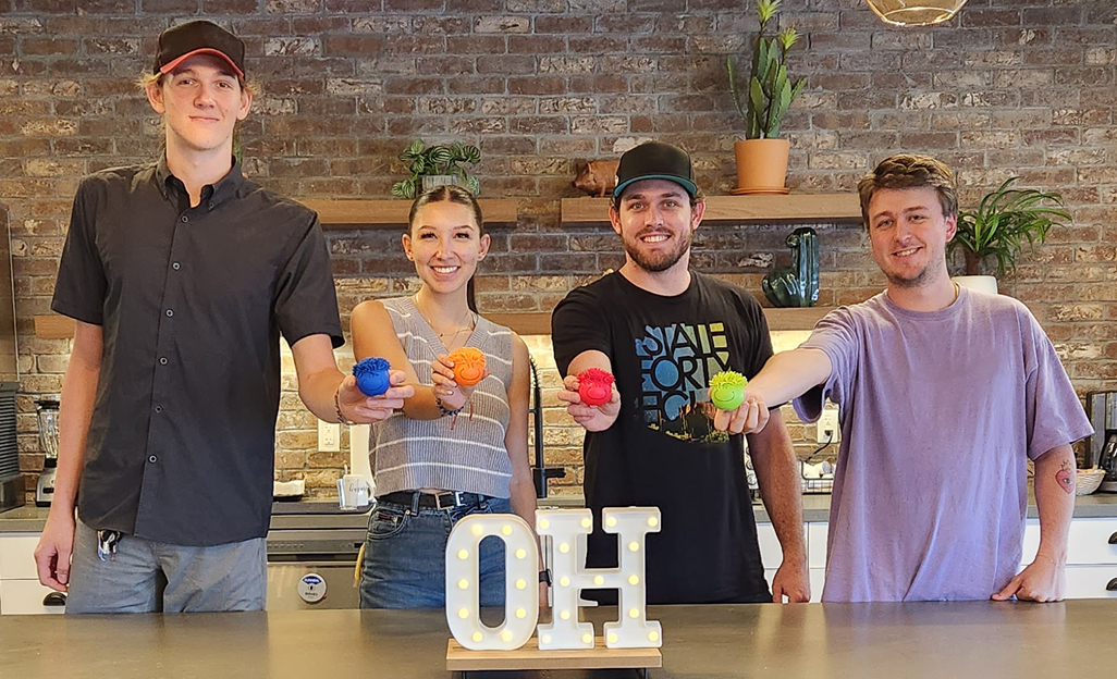 Four team members holding colorful stress relievers in front of "O.H." sign.