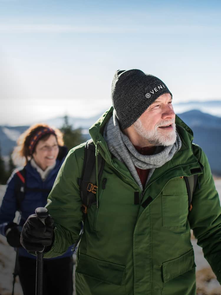 Man wearing coat and beanie with camera and person behind him