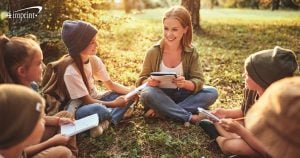 Group of kids and one adult sitting in discussion circle outside with notebooks and pens