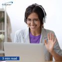 A woman in headphones waves at a computer screen.