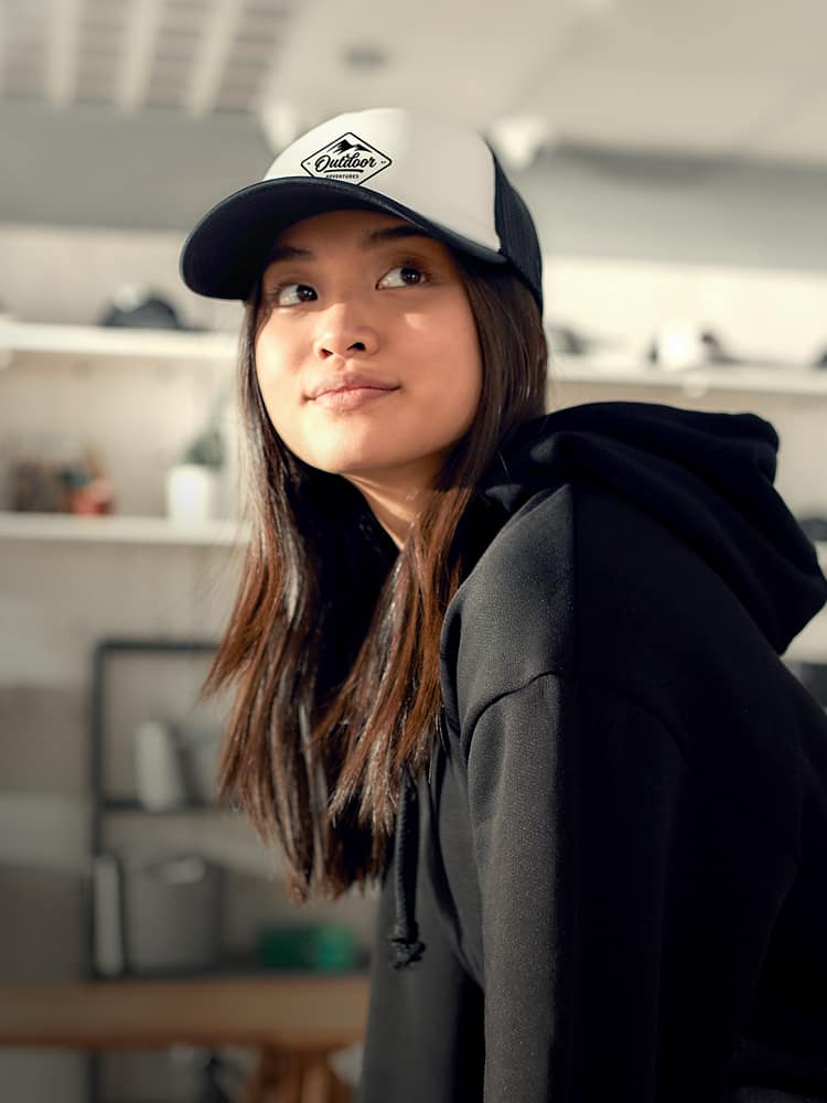 Young woman wearing a hooded sweatshirt and a trucker hat with a logo.