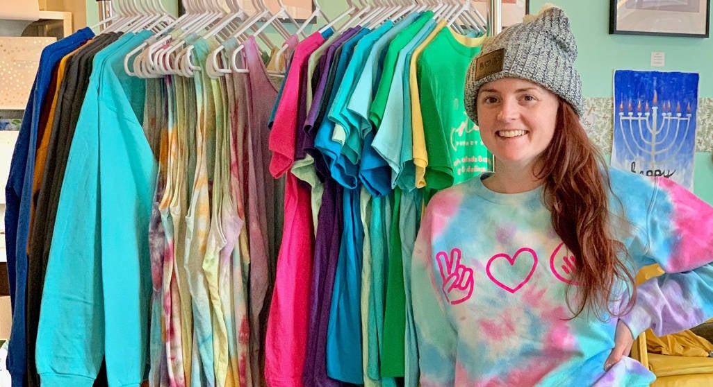 Woman in branded sweatshirt and hat standing next to rack of branded merchandise she is selling