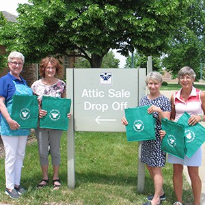 Four volunteers holding their green canvas grocery totes, posing by their drop-off sign