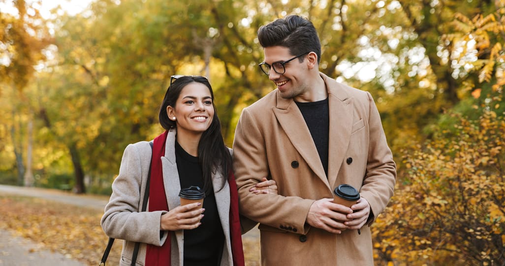 A man and woman walking through the park in the Fall with cups of coffee in their hands